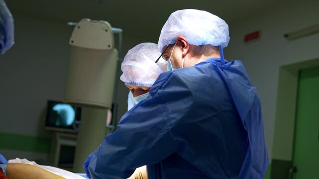 Surgeon with glasses working in operating room. Two medical workers in blue gowns face toward a patient table near a blurred monitor screen.