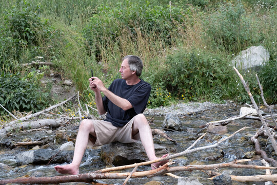 Middle aged white man sits on a rock in a small rocky stream at the base of Stewart Falls, Mount Timpanogos,  Solo traveler taking a photo of nature in the mountains on mobile phone in a river.