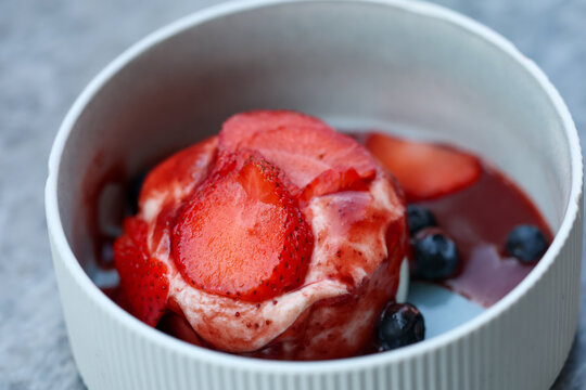 Creamy dessert topped with fresh strawberries and blueberries in red sauce, served in a white bowl.
