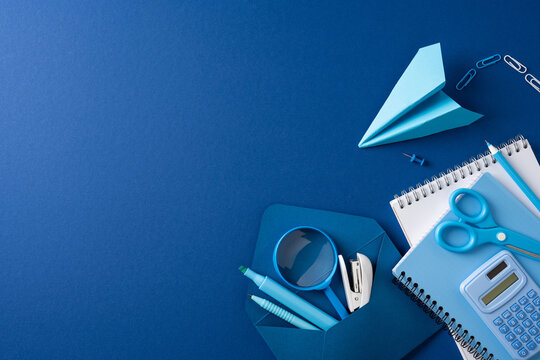Back to school flat lay with blue stationery, calculator, scissors and paper airplane on blue background
