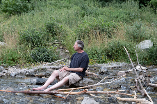 Caucasian man sits on a rock with feet in the cold water at the base of Stewart Falls.  Man sites by a stream having a quiet moment of peace in nature.