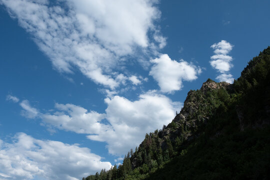 Sunlight on the top of Mt Timpanogos on a summer day with deep blue clouds and fluffy white clouds near Sundance, Utah.  Tall rocky mountain covered with pine trees.