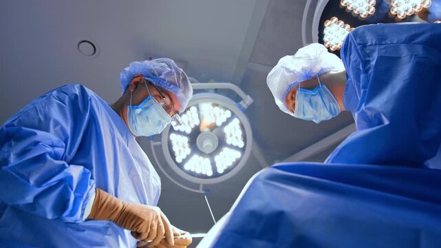 Low angle view of surgeons looking down during operation. Two medical professionals in blue scrubs work beneath a circular ceiling lamp inside a clinic.
