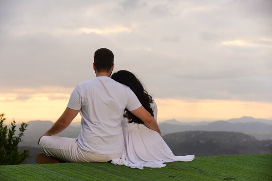 romantic couple sitting on hilltop enjoying mountain vista view at sunset, man and woman dressed in white sitting on green grass looking at horizon, lovers embracing at scenic mountain viewpoint dusk 