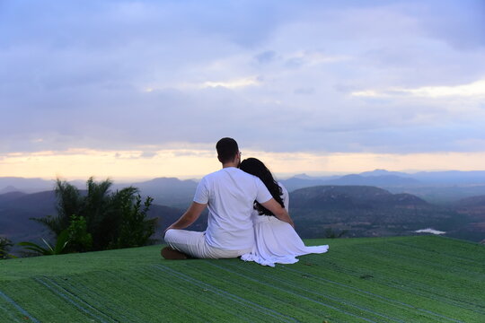 romantic couple sitting on hilltop enjoying mountain vista view at sunset, man and woman dressed in white sitting on green grass looking at horizon, lovers embracing at scenic mountain viewpoint dusk 
