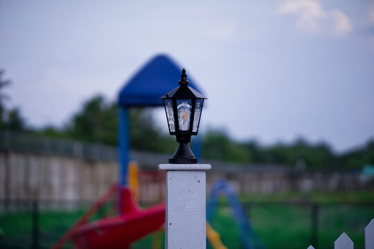 A decorative outdoor lantern light fixture mounted on a white wooden fence post in a park setting