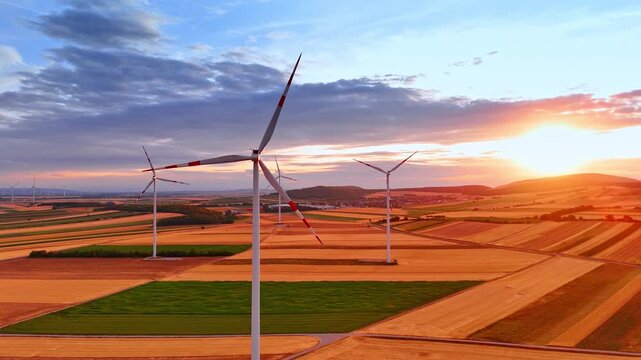 Wind turbines in fields at sunset. Bright sunlight glows on the horizon over golden agricultural rolling hills with modern electricity generators.