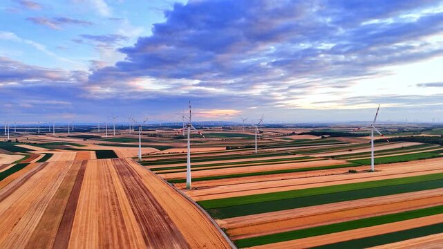 Aerial view of multiple wind turbines. Numerous white structures are scattered across striped green and yellow farm fields under a cloudy sky.