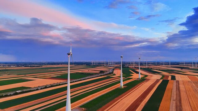 Wind turbines along fields at sunset. A row of tall electricity generators stretches across colorful striped rural lands under pink clouds.