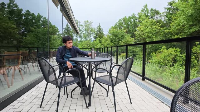 Man relaxes at outdoor table, sipping drink. Garden views frame his calm, contemplative moment. Glass walls reflect trees and sky. Perfect for tranquil, modern, urban scenes