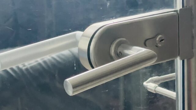 Closeup metal door handle on glass, brushed stainless lever with visible keyhole and hinge, sleek commercial entrance capturing reflections Taken on mobile device.