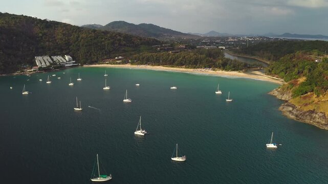 Yachts resting in the clear blue water of Nai Harn Bay. Pristine sand beach curving along the lush tropical landscape and green hills of Phuket island, travel Thailand. Aerial view drone panorama