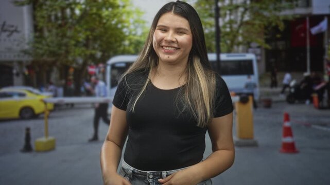 Young woman smiling with hands on chest and flipping hair, wearing black top and denim jeans on street near taxi and cones; youthful happiness.