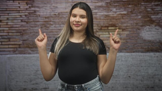 Hispanic young woman pointing index fingers with both hands visible, smiling slightly, wearing a black t shirt and denim jeans in front of a brick building; playful confidence.