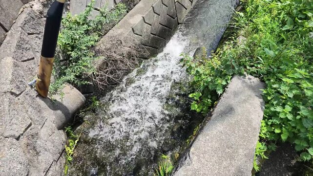 Water rushes through cracked concrete. Tires lie beside the streambed. Green foliage grows wild around the runoff. Flow creates white foam against stone edges