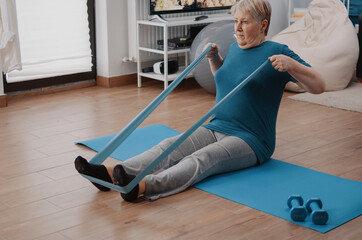Fototapeta premium Elderly woman exercises with resistance band using an elastic band for stretching, muscle training and gentle rehabilitation during a home fitness routine.