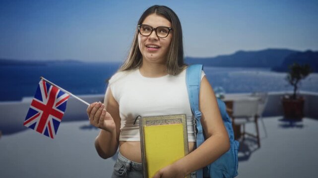 Young woman holds uk flag and textbooks with visible hand, wearing glasses and a backpack strap on shoulder in a studio; aspiration.