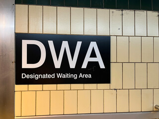 Obraz premium Black DWA Designated Waiting Area sign with white text mounted on yellow subway tile wall in subway transit station, Toronto July 2026