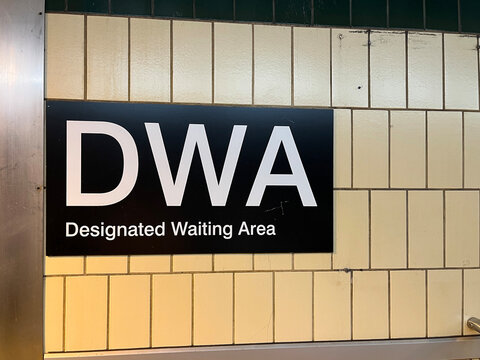 Black DWA Designated Waiting Area sign with white text mounted on yellow subway tile wall in subway transit station, Toronto July 2026