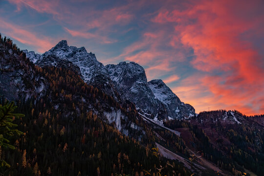 Alpine landscape with snow-covered mountain peaks and autumn coloured forested hills stands against dramatic clouds glowing with fiery red and pink hues during a beautiful sunset. Austria 