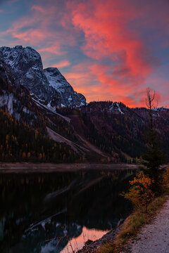The dramatic clouds glowing with fiery red and pink hues over a snowy towering mountain peaks and calm water of Gosau lake (Upper Gosausee). Austria 
