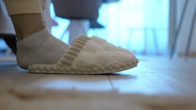 Woman putting on cozy slippers at home and walking away. Low angle view of female feet wearing socks sliding into comfortable and warm plush slippers on a wooden floor, and then walking away