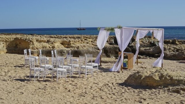 Beautiful wedding arch with white fabric curtains and guest chairs standing on a sandy beach by the sea, prepared for a romantic destination ceremony on a windy day with a boat in the background