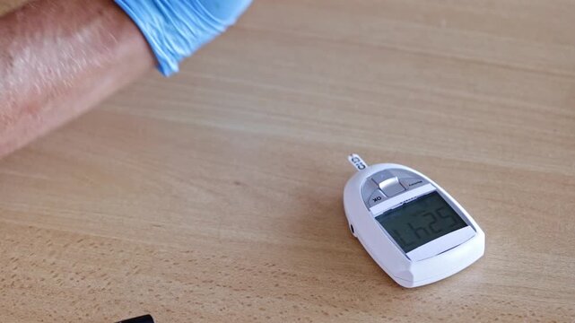 Medical professional wearing blue gloves using a glucometer to measure a patient's blood glucose level, placing a drop of blood on the test strip to monitor for diabetes or hypoglycemia
