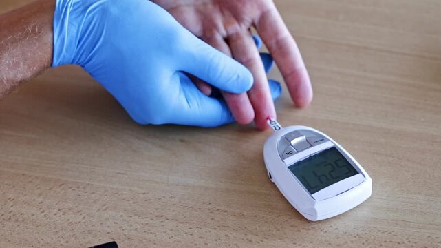 Medical professional wearing blue sterile gloves is performing a glucose test on a patient's finger, using a portable glucometer to measure the blood sugar level for diabetes monitoring