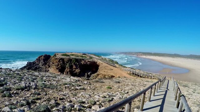 First person perspective walking along a scenic wooden boardwalk leading to the beautiful and vast praia da bordeira on the vicentina coast, a popular surfing spot in the algarve, portugal