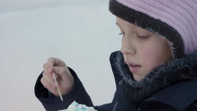 Young girl in a knit hat and scarf eats a sweet dessert from a small wooden spoon outdoors in winter