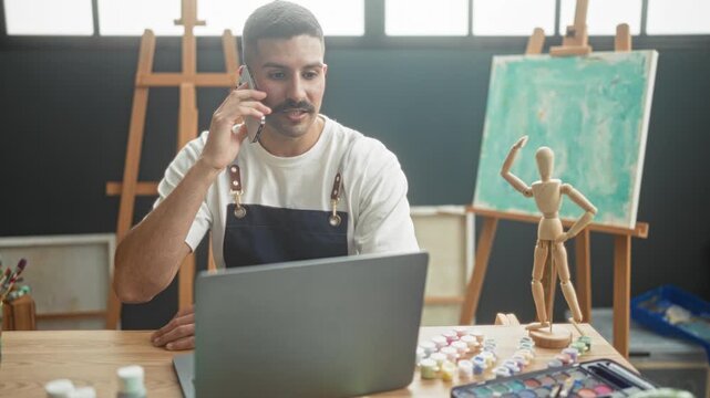 Man painter in studio wearing apron holds smartphone to ear while working at laptop beside paints and wooden mannequin; creative focused.