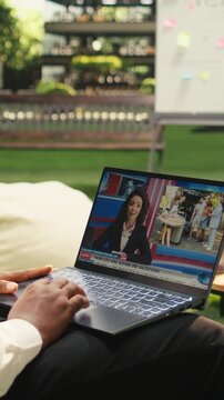 Vertical screen: Black businessman watching laptop news report about artificial intelligence, online news of current affairs in informal workplace, video streaming, outdoor corporate culture.