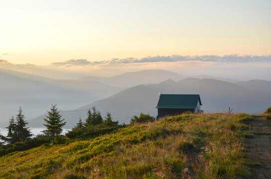 Solitary cabin sits on a grassy mountain ridge overlooking a vast, misty valley. Sunbeams pierce through distant clouds over rolling hills and scattered evergreen trees at dawn