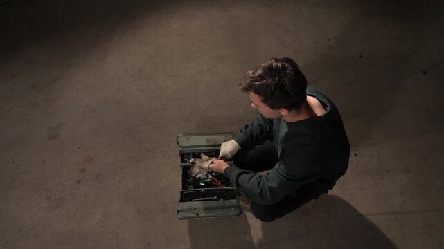 High angle view mechanic man squatting on the concrete floor, wearing protective gloves and reaching inside an open metal toolbox to find a needed tool for repair or maintenance work