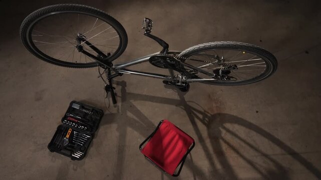 High angle view bicycle upside down on a concrete garage floor, next to an open toolkit with various repair instruments and a red folding stool, ready for maintenance