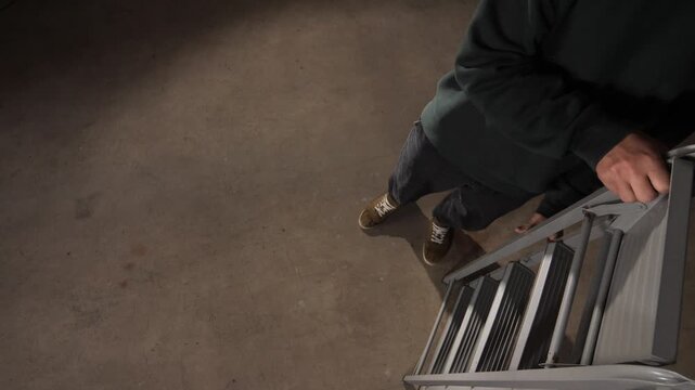 High angle view person's hands and foot are visible while stepping onto a gray metal folding step ladder, preparing to work or reach an elevated area within an indoor setting