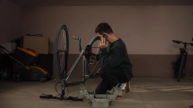 Young man crouching in a home garage, performing maintenance on a bicycle with tools while discussing the repair on a mobile phone for guidance