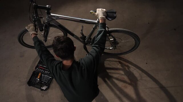 High angle view person wearing gloves on the floor of a garage, actively performing maintenance on a bicycle with a toolbox nearby, emphasizing diy culture and cycling passion