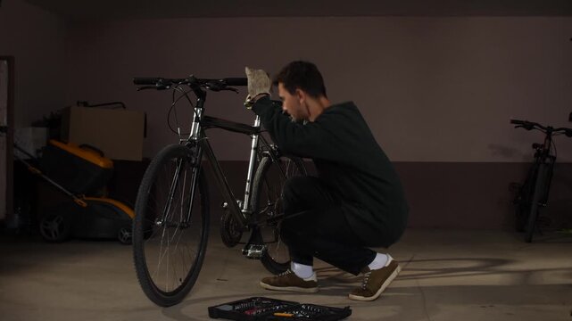 Man wearing work gloves crouching and carefully maintaining a bicycle in a dimly lit garage or workshop, with a tool kit open on the floor and other equipment in the background