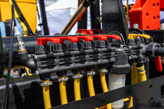 Agricultural sprayer control valve manifold block section. Modern farm machinery liquid distribution mechanism with red levers on black chassis frame and industrial equipment technical details.