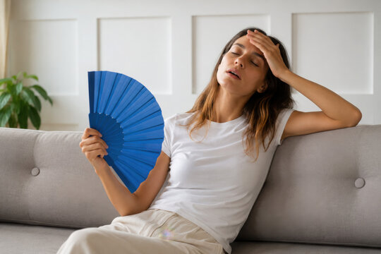 Woman at home suffering from overheating, sitting on sofa in living room holding hand paper fan for cooling down. Tired young female on couch with sick expression, overwhelmed by summer heat or fever