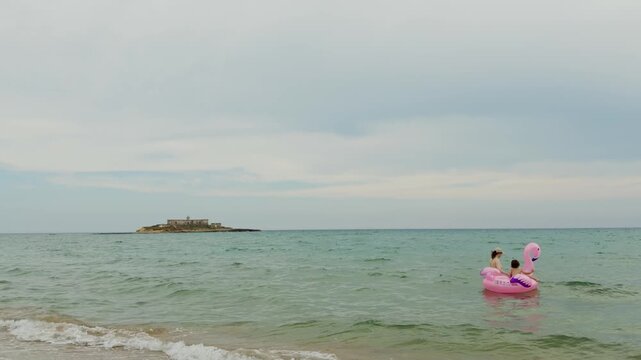 Mother and toddler enjoying a summer afternoon by the Mediterranean Sea with a pink flamingo float and Isola delle Correnti in the background.