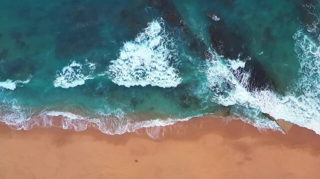 Drone flies above turquoise Atlantic waves breaking onto a wide sandy beach, revealing vivid coastal textures and white sea foam.