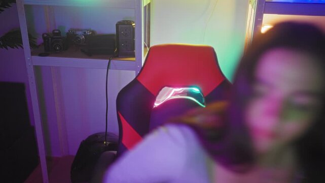 Woman leans forward from red gaming chair toward camera with visible necklace in a streaming studio lit by neon lights; friendly warmth.