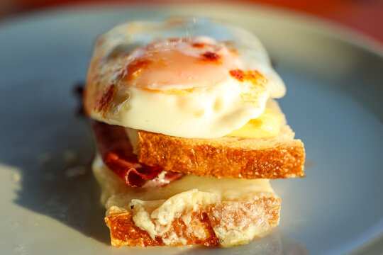 A close-up of a Croque Madame sandwich with melted cheese, ham, and a fried egg on toasted bread in warm sunlight.