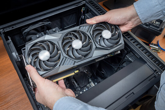 A technician holds a powerful gaming graphics card above an open personal computer case. Expensive computer components. A powerful and expensive PC with scarce components