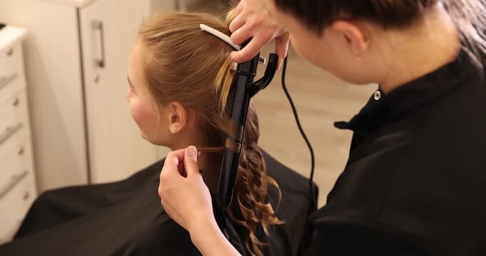 Hairdresser styling woman's hair with curling iron in beauty salon, closeup