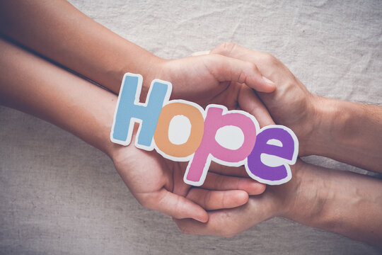 Hope, hands, child, adult, support, help, trust concept showing an adult and child holding the word hope together, symbolizing care, community, charity and a bright future for all.
