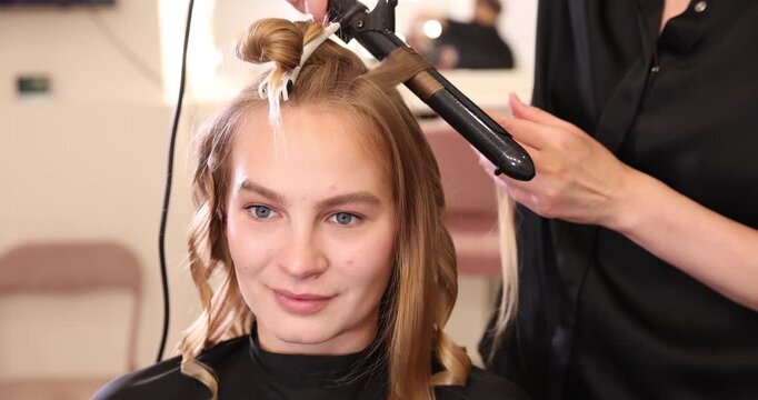 Hairdresser styling woman's hair with curling iron in beauty salon, closeup